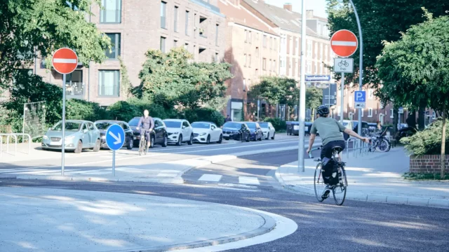 Kreisverkehr an der Hufnerstraße in Hamburg mit Einmündung in die Poppenhusenstraße (Protected Bike Lane); eine Radfahrer:in im T-Shirt biegt bei gutem Wetter rechts ab. © BVM / Robin Lindner Kreisverkehr an der Hufnerstraße in Hamburg mit Einmündung in die Poppenhusenstraße; eine Radfahrer:in im T-Shirt biegt bei Sonnenschein rechts in die Protected Bike Lane ab.