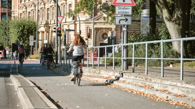 Protected Bike Lane am Stephansplatz in Hamburg: Eine Radfahrerin fährt bei gutem Wetter, weiter hinten warten Radfahrende an einer roten Ampel. © BVM / Robin Lindner Stephansplatz in Hamburg mit Protected Bike Lane; eine Radfahrerin von hinten, weiter hinten warten Radfahrende an roter Ampel.